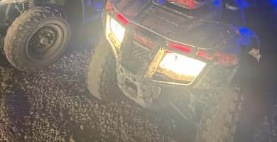 A muddy quad with its headlights on at night on a gravel trail