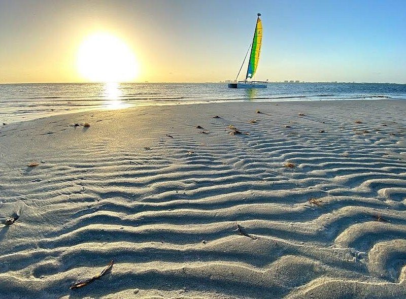 Rippled sand at the water's edge with the sun low and a small sailboat offshore