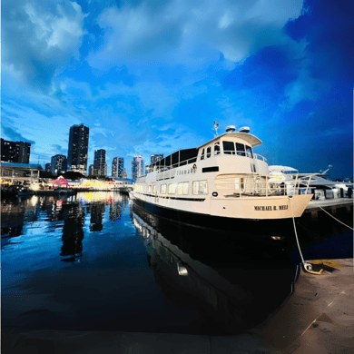 A two-deck cruise boat moored at a downtown dock at dusk, towers behind