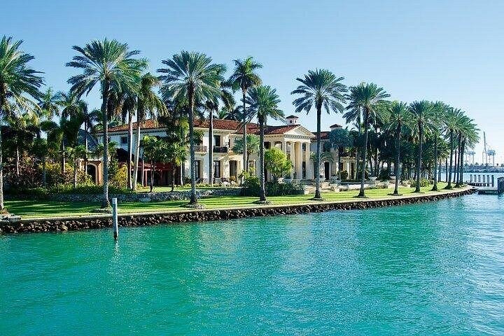 A Mediterranean-style mansion behind palms and a sea wall on turquoise water